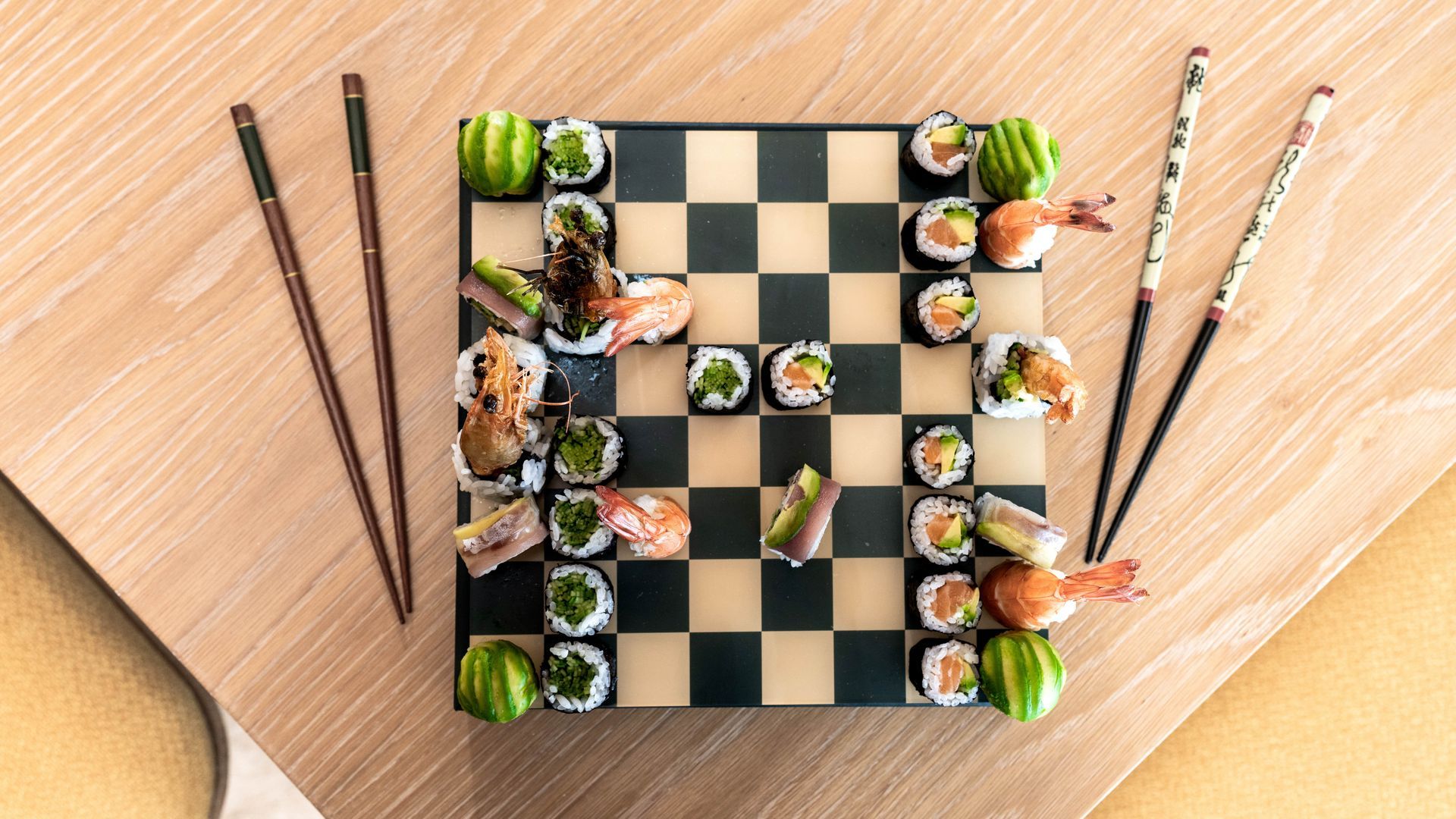 A chess board filled with sushi and chopsticks on a wooden table.