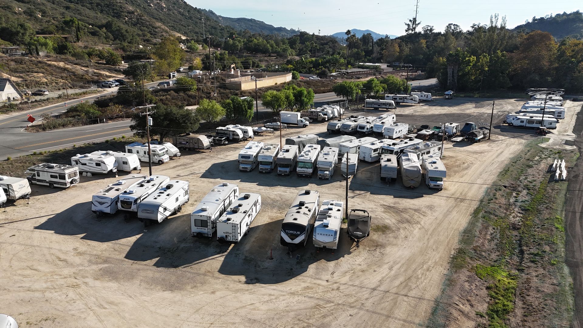 RV campers parked in a row on a gravel lot under a partly cloudy sky. Trees in the background.
