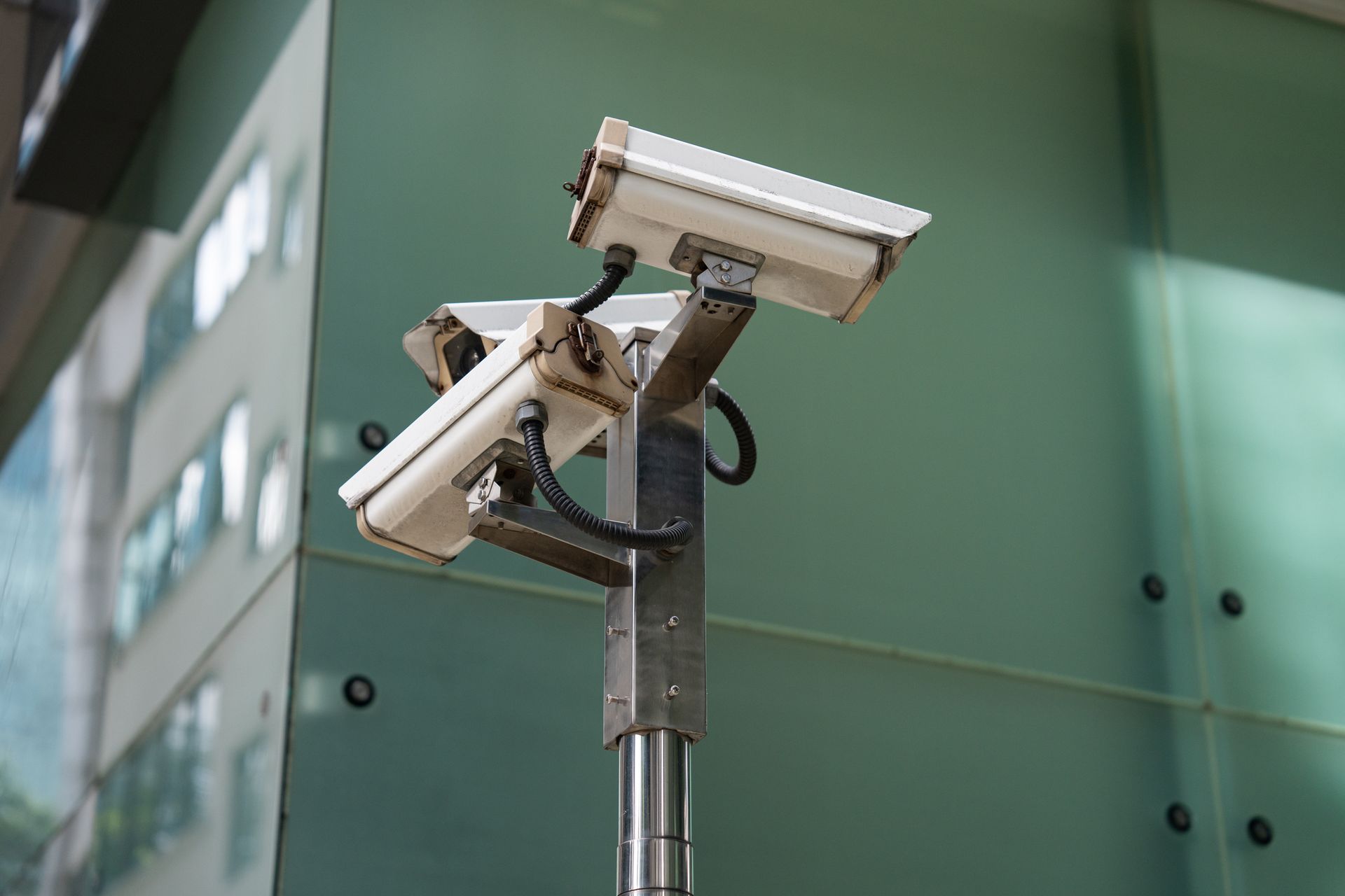 Two white security cameras on a metal pole, mounted in front of a glass building.