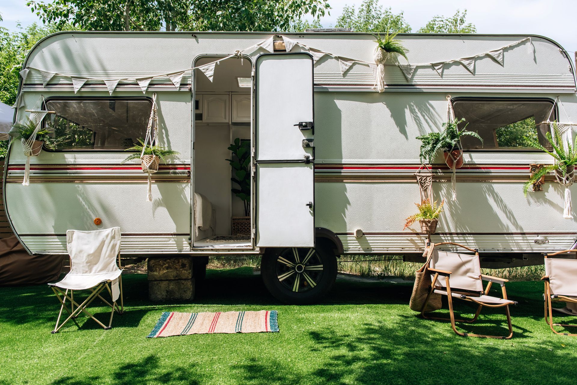 Vintage trailer with open door on green lawn, decorated with plants and bunting, with chairs.