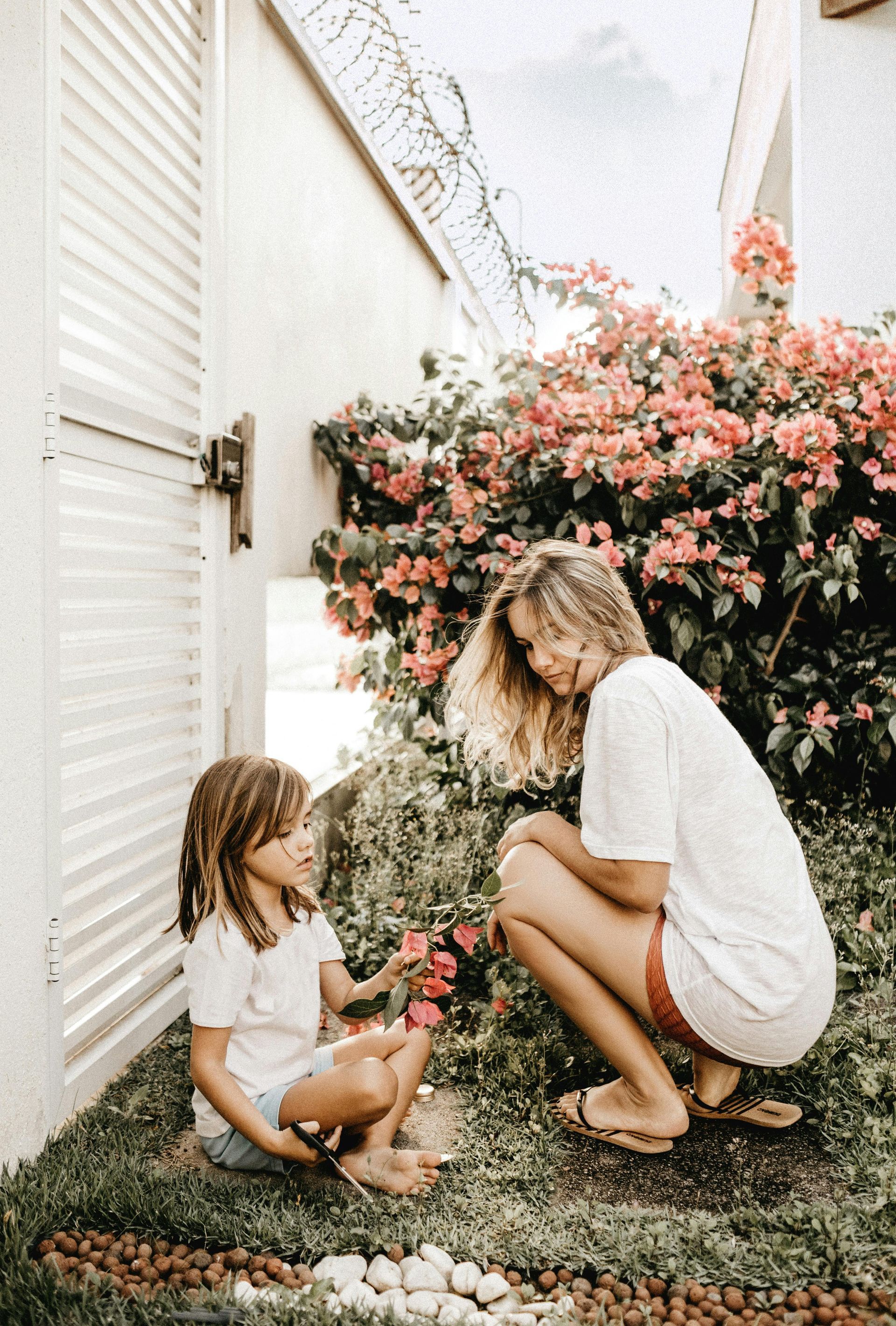 Woman and child kneeling in garden, examining pink flowers.