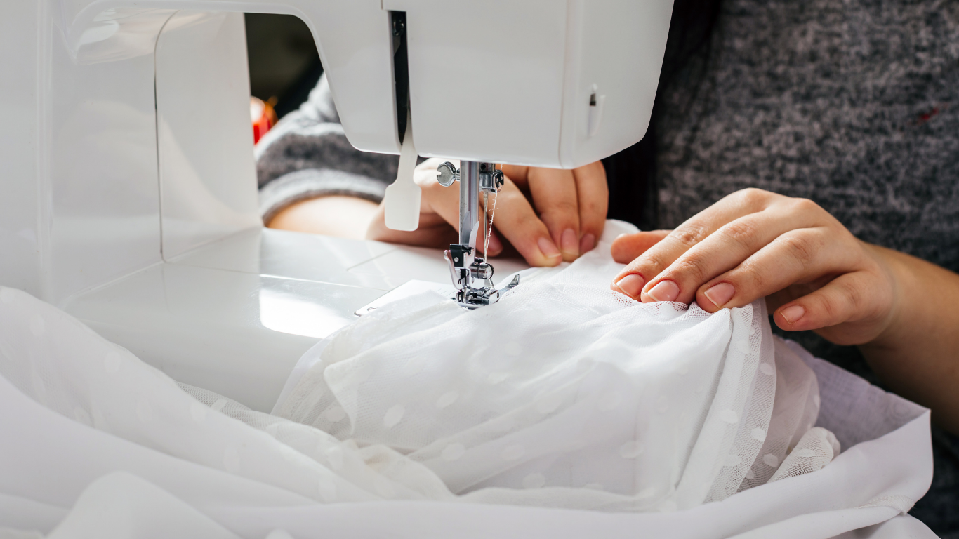 Woman with glasses sewing at a table with white fabric. Sewing machine, lamp, and light-colored wall.