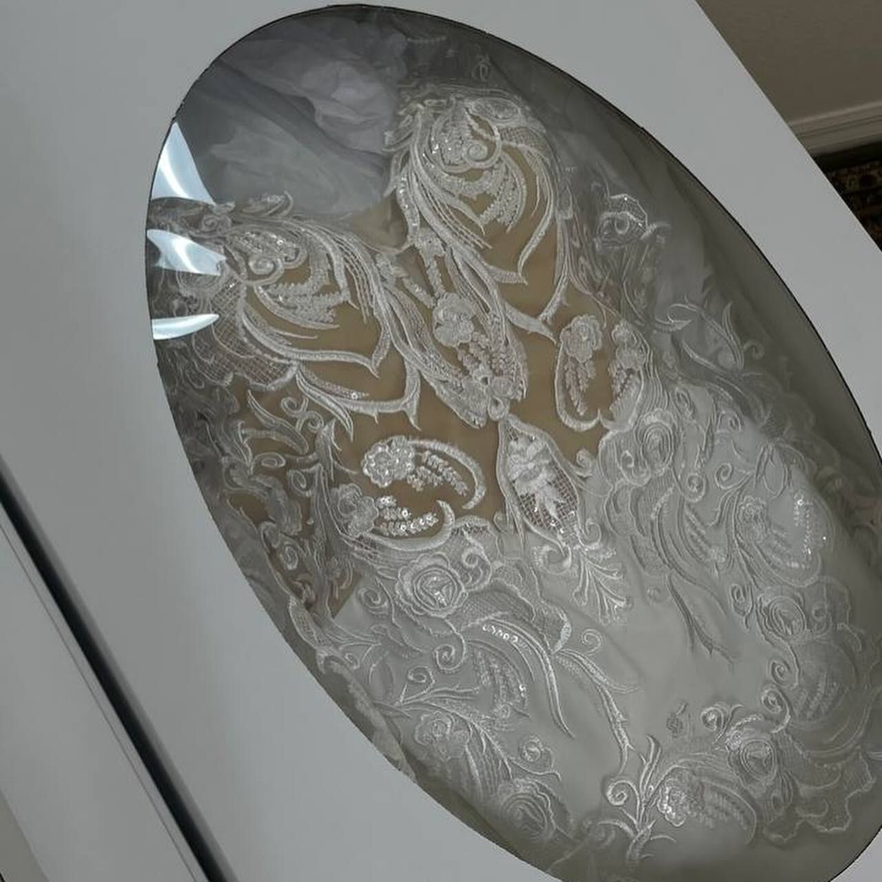 White wedding dress with intricate lace details in an oval display window.