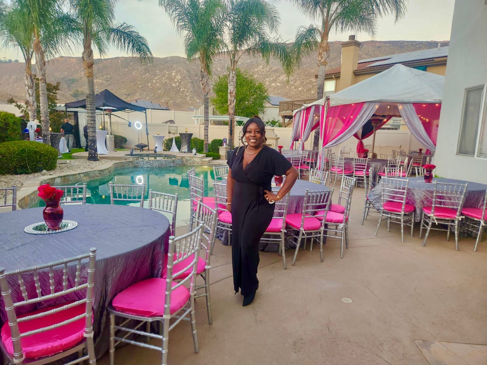 Woman in black jumpsuit poses at an outdoor party with pool, pink and gray decor.