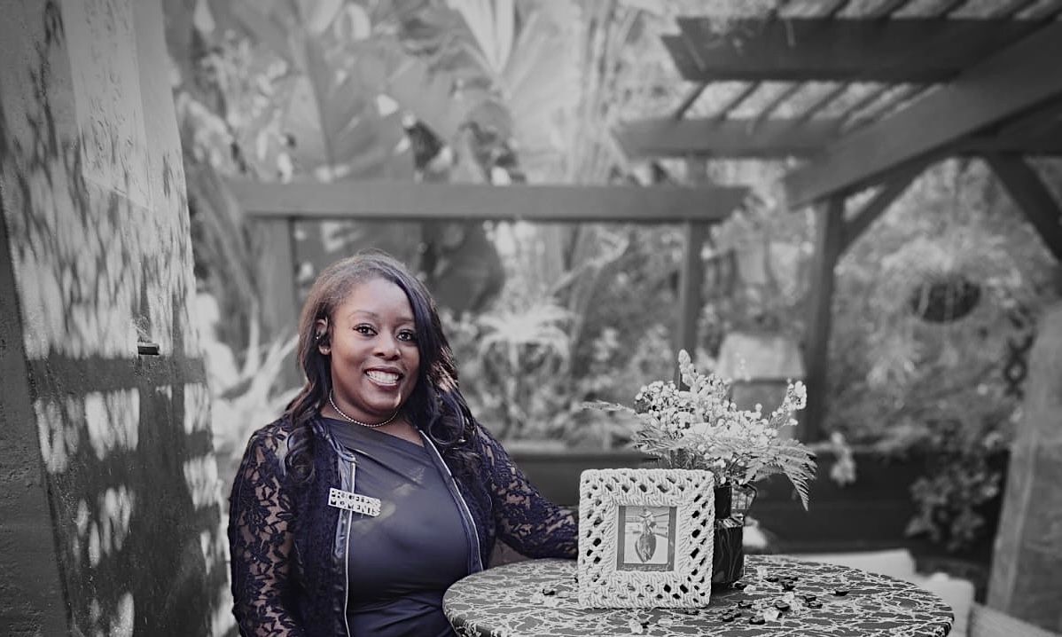 Woman seated at a table with a decorative frame in a garden setting, smiling.