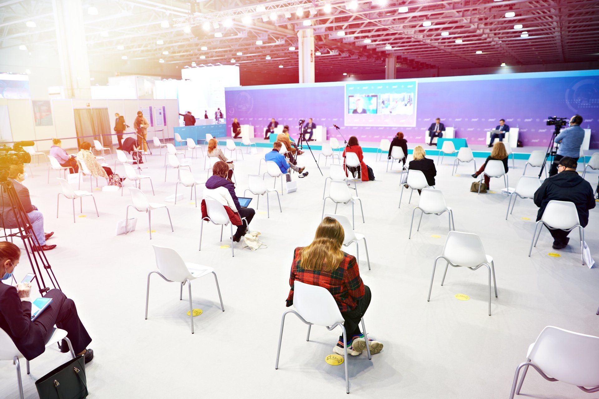 People seated in a large, bright hall, spaced apart, possibly an event or waiting area.