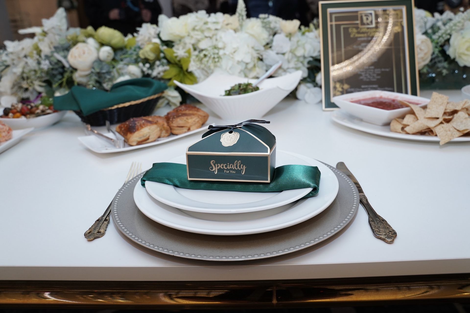 Formal table setting with white, green and gold accents; small gift box on a plate.