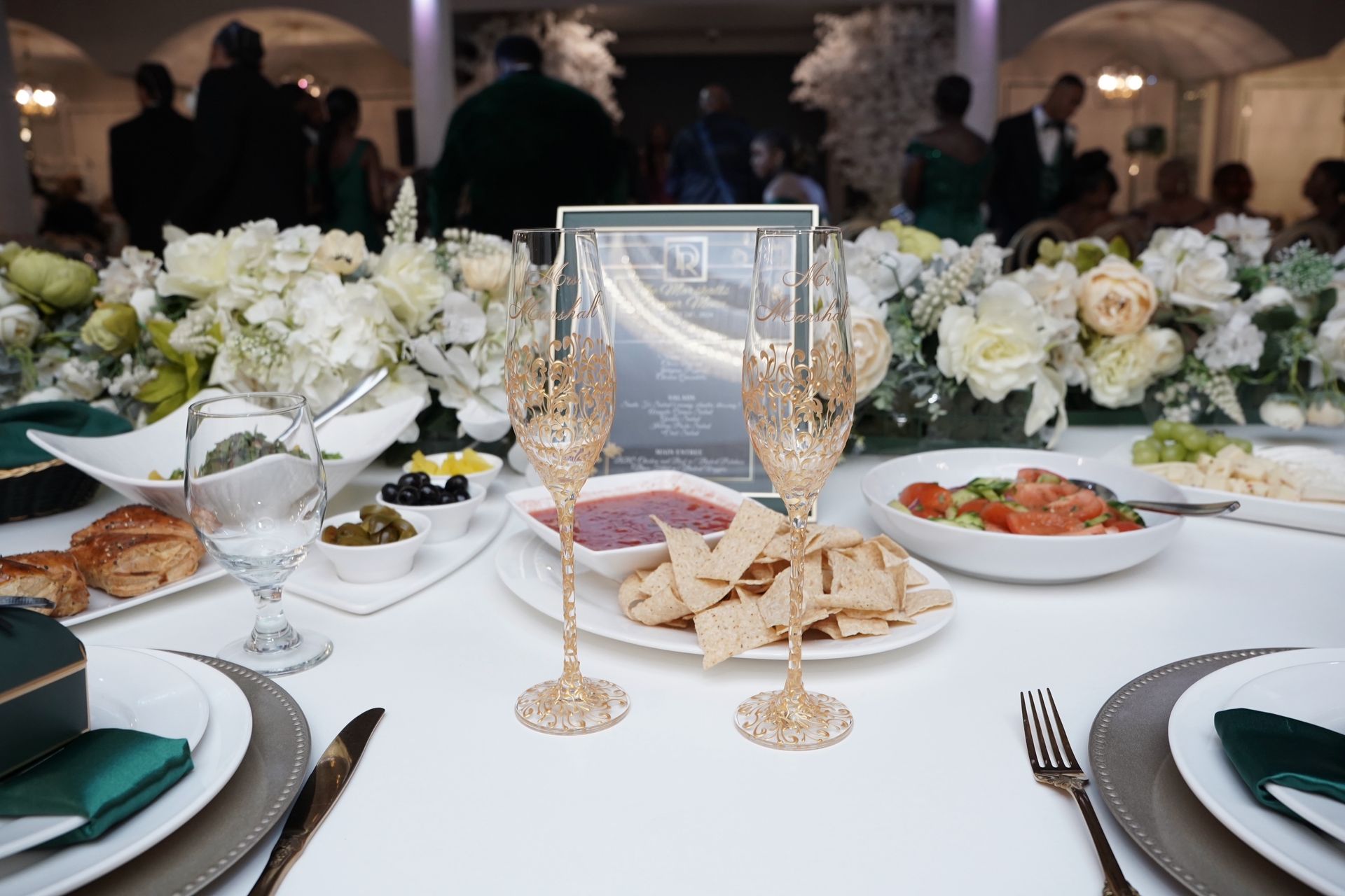 Formal dinner table setting with food, flowers, and champagne flutes; guests in background.