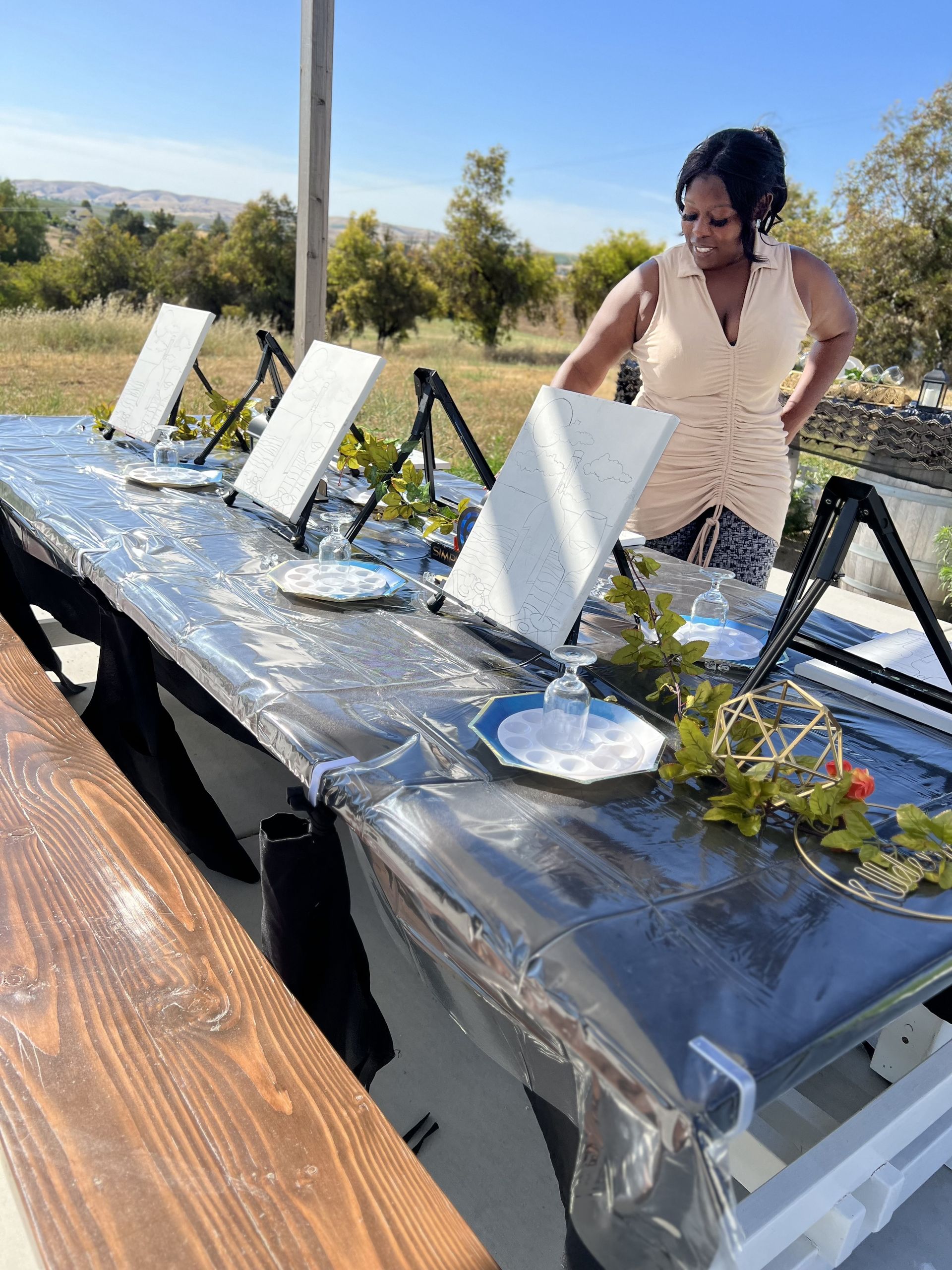 Woman at an outdoor art class, setting up canvases and supplies on a long table covered in black plastic.