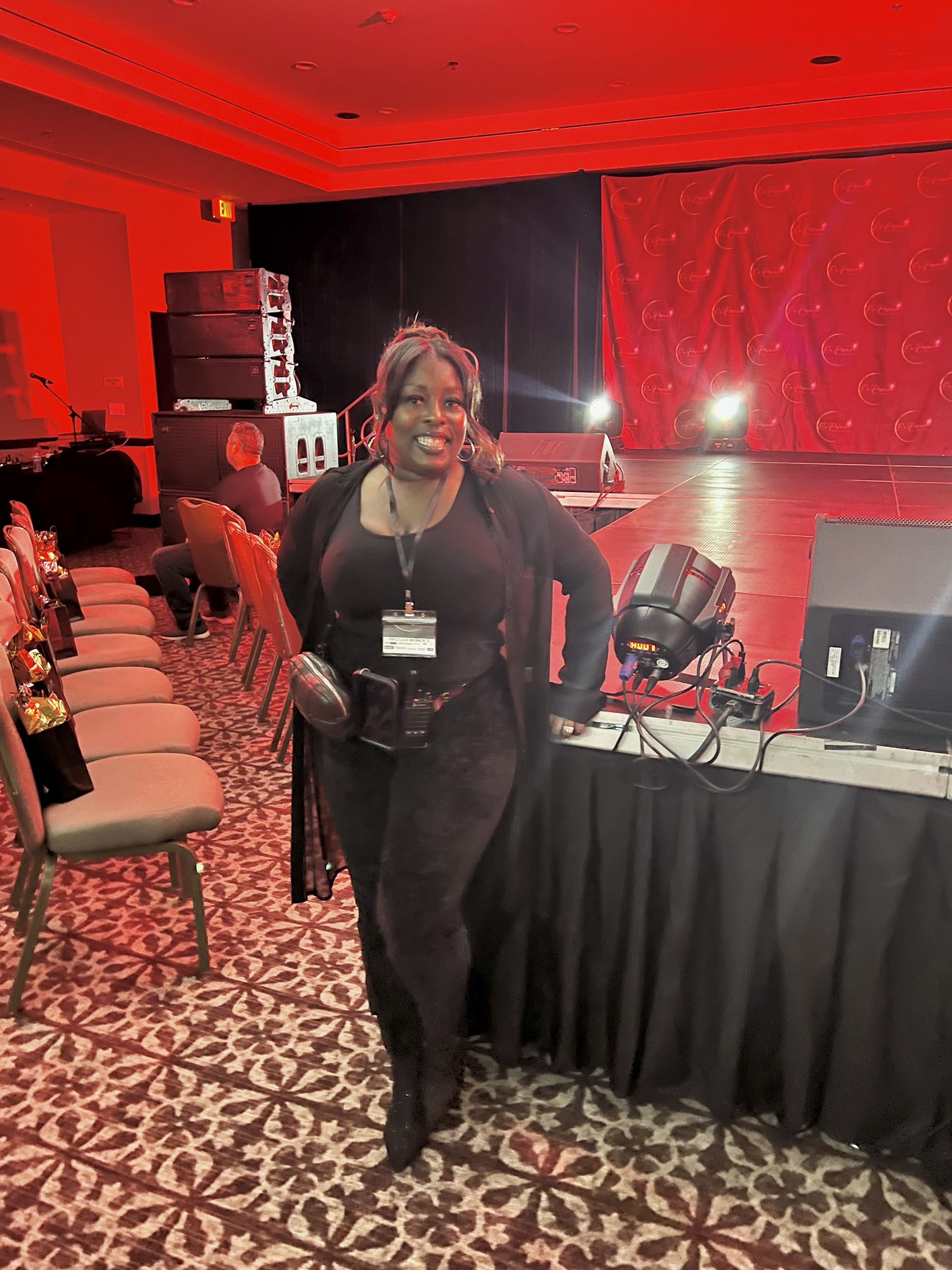 Woman in black outfit stands near sound equipment on a stage. Red lighting fills the room.