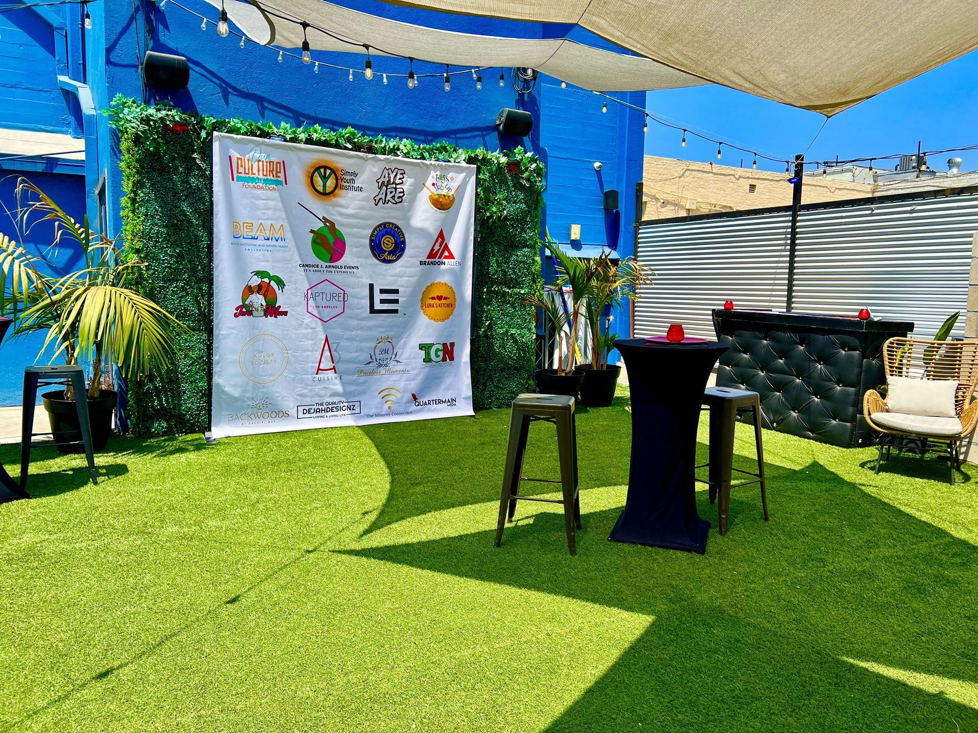 Outdoor event space with logo banner, green wall, artificial turf, and bar seating.