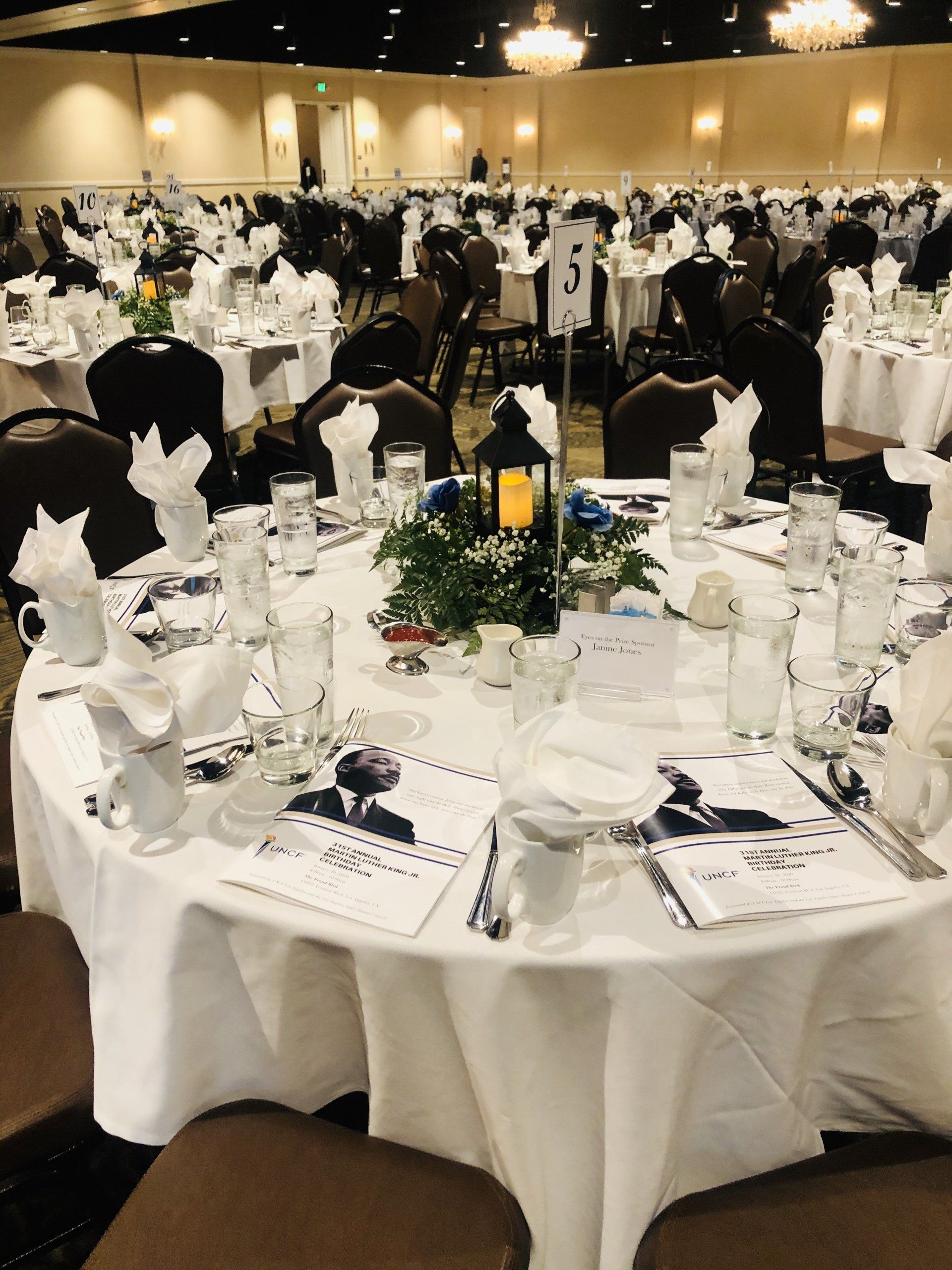 Round table set for an event, white linens, centerpieces, place settings, and chairs.