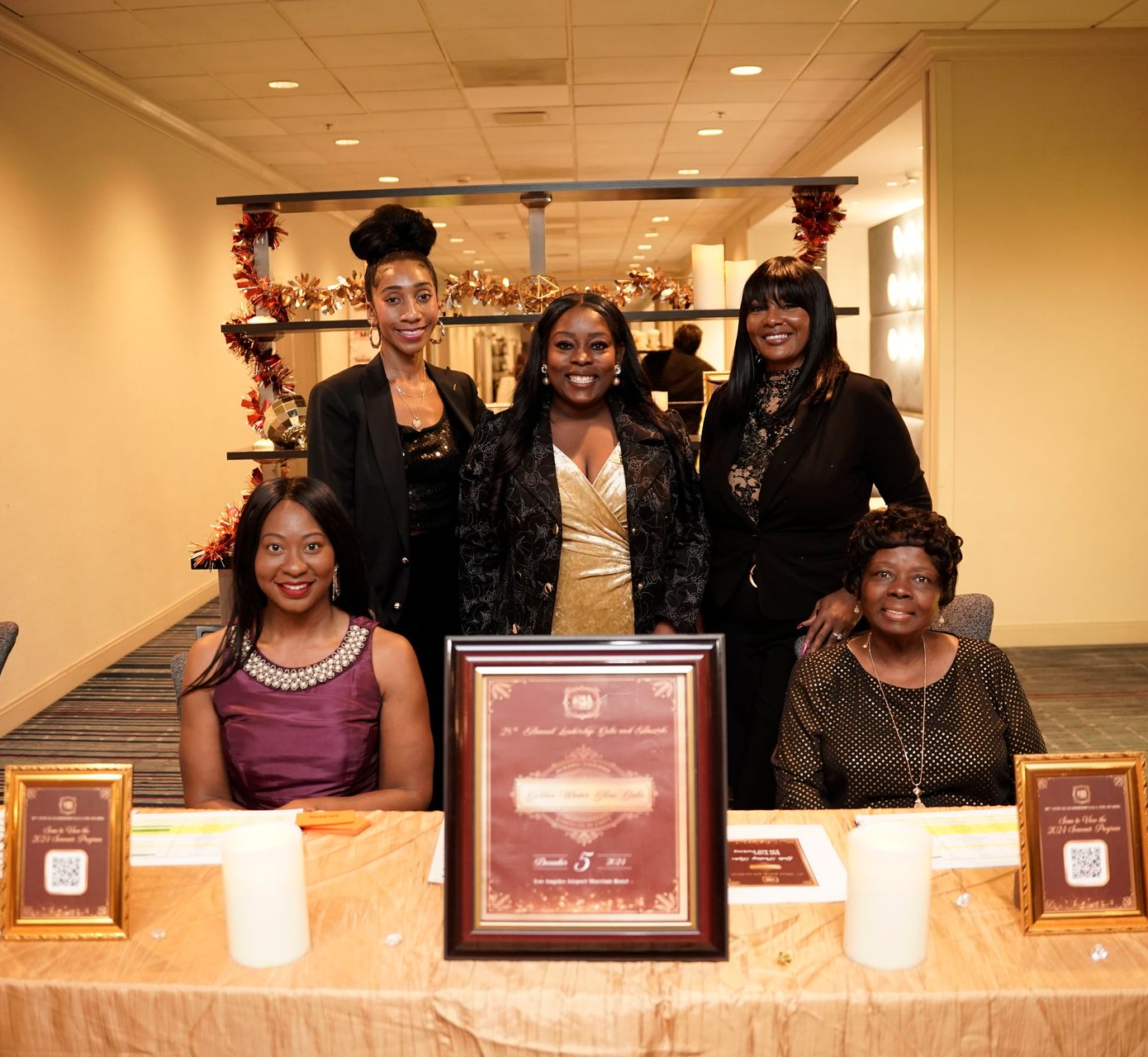 Five people at a table; a framed award sits in front. Warm lighting, a gold tablecloth, and autumn decor are present.