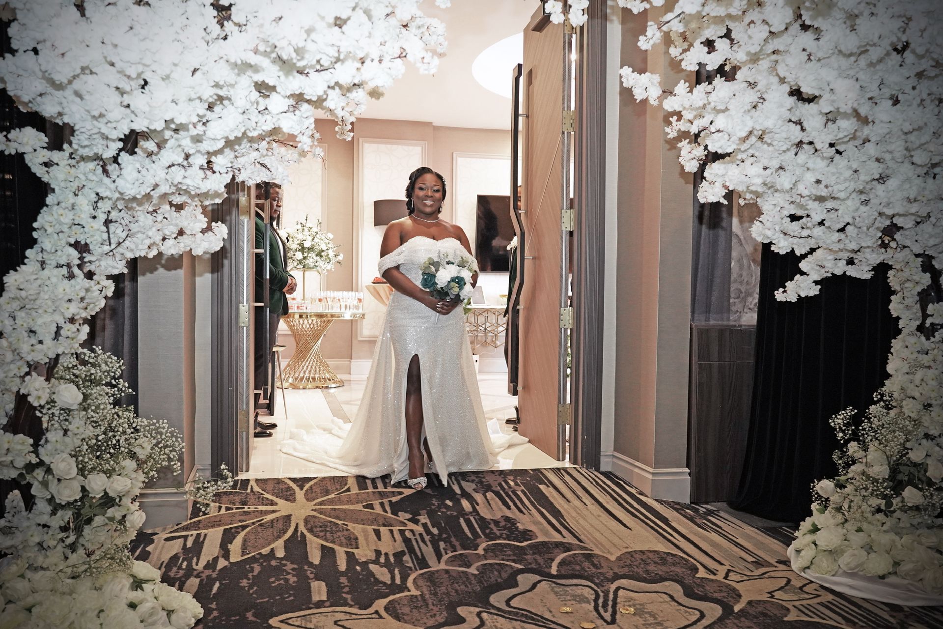 Bride in a white gown with a high slit, holding a bouquet, standing in a doorway decorated with white flowers.
