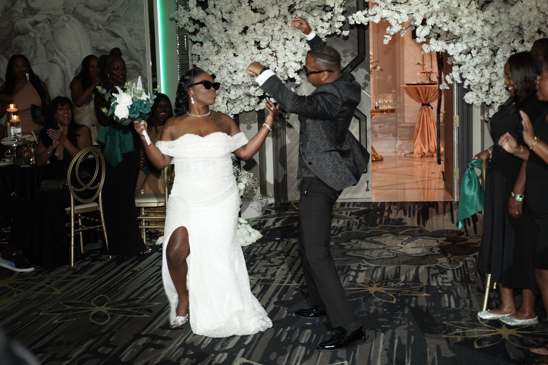 Newlyweds dancing at a wedding reception. Woman in white dress and man in suit wear sunglasses, celebrating. White floral archway.