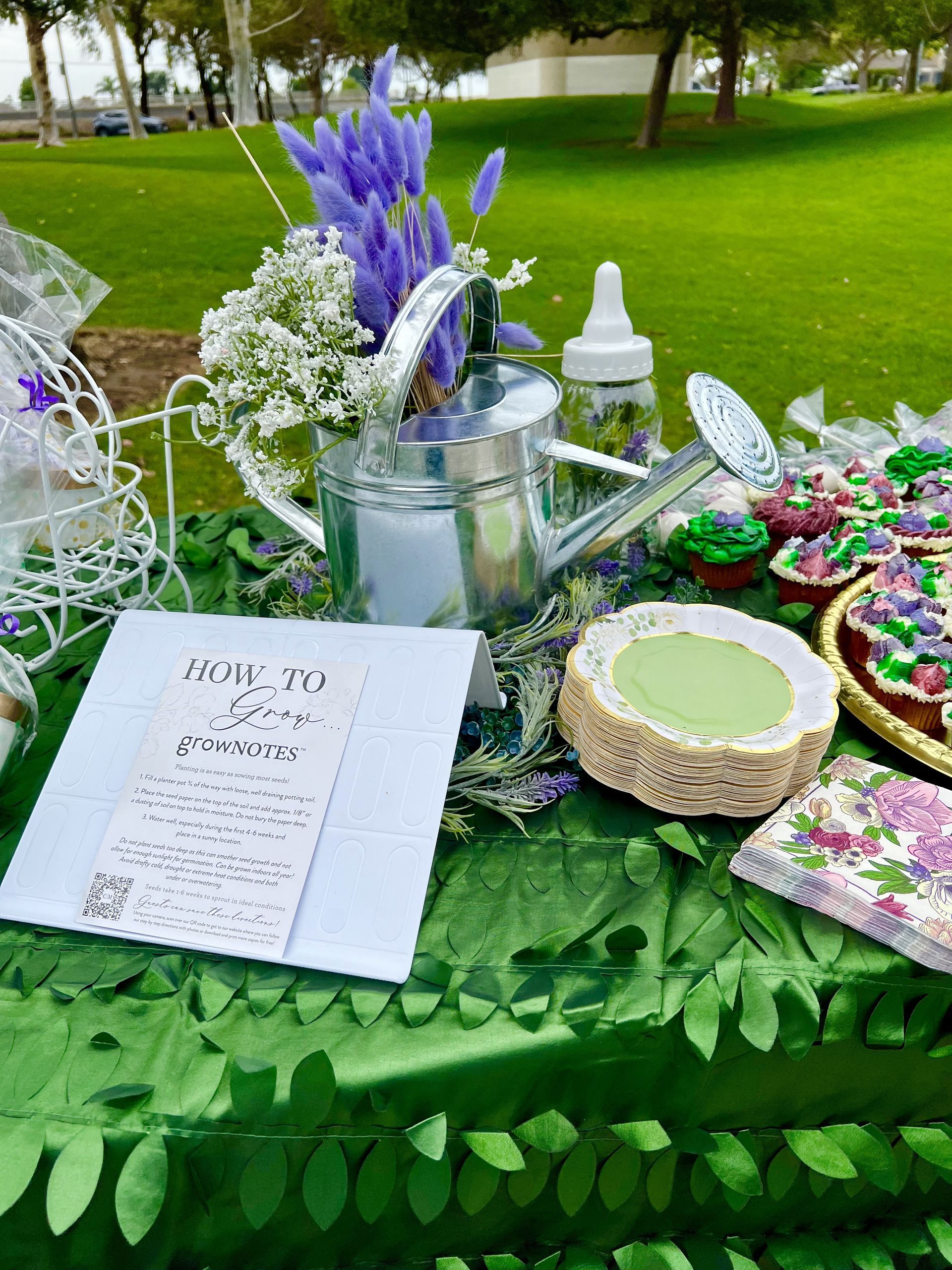 A table decorated with lavender flowers, treats, and a watering can, set in a park.