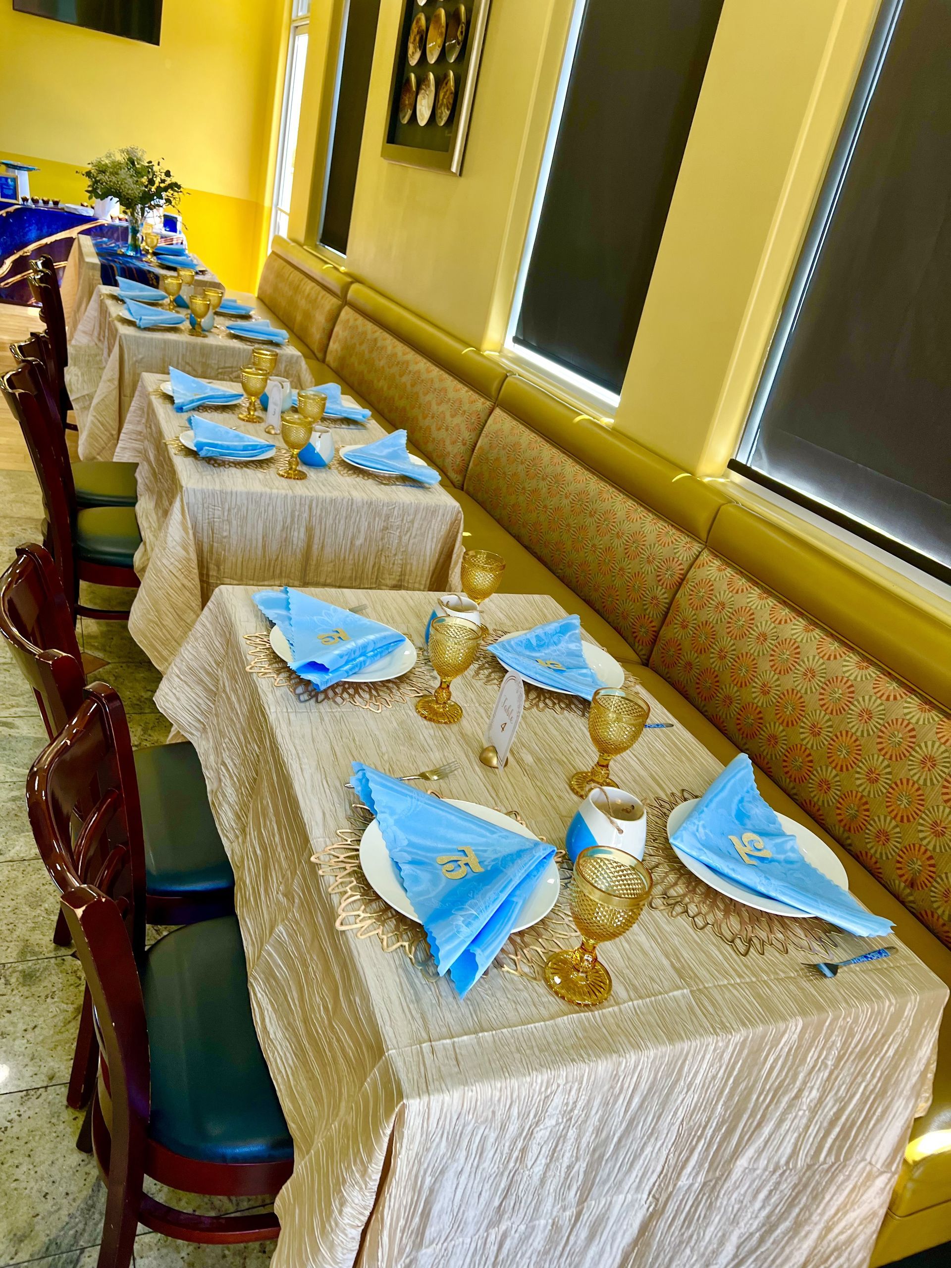 Tables set for a party, with gold tablecloths, blue napkins, and goblets, arranged along a cushioned bench in a restaurant.
