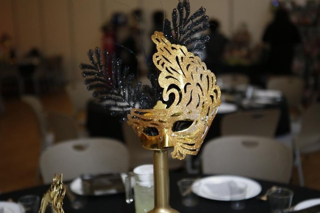 Gold mask centerpiece with black feathers on a table set for an event.