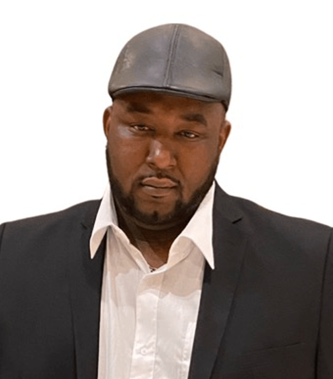 Man in a suit and hat, looking down, slight smile.