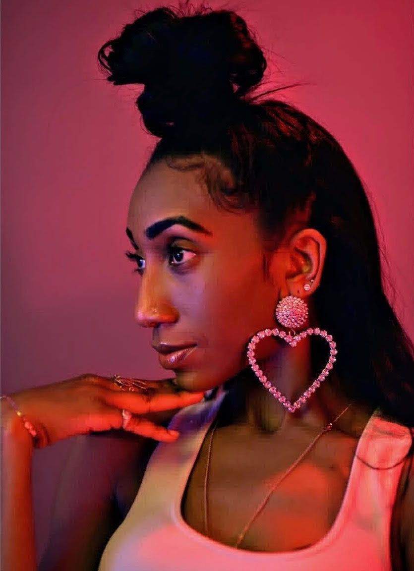 Woman with topknot and large heart earrings, pink lighting.