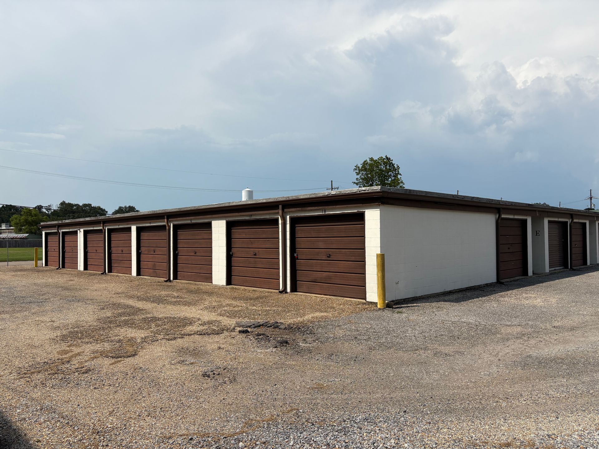 Storage units with brown doors, gravel lot, under a cloudy sky.