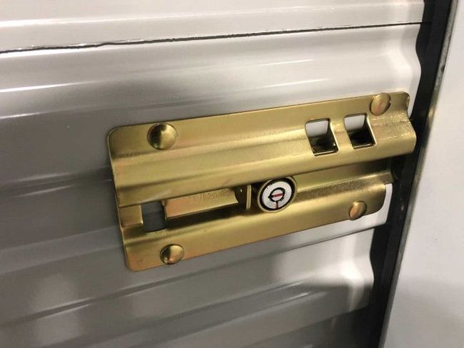 A golden metal storage unit latch with a keyhole, bolted to a corrugated gray door.
