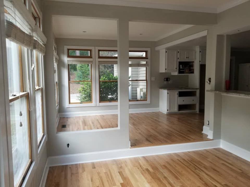 Interior view of room with hardwood floors, windows, and partial walls; kitchen visible in the back.