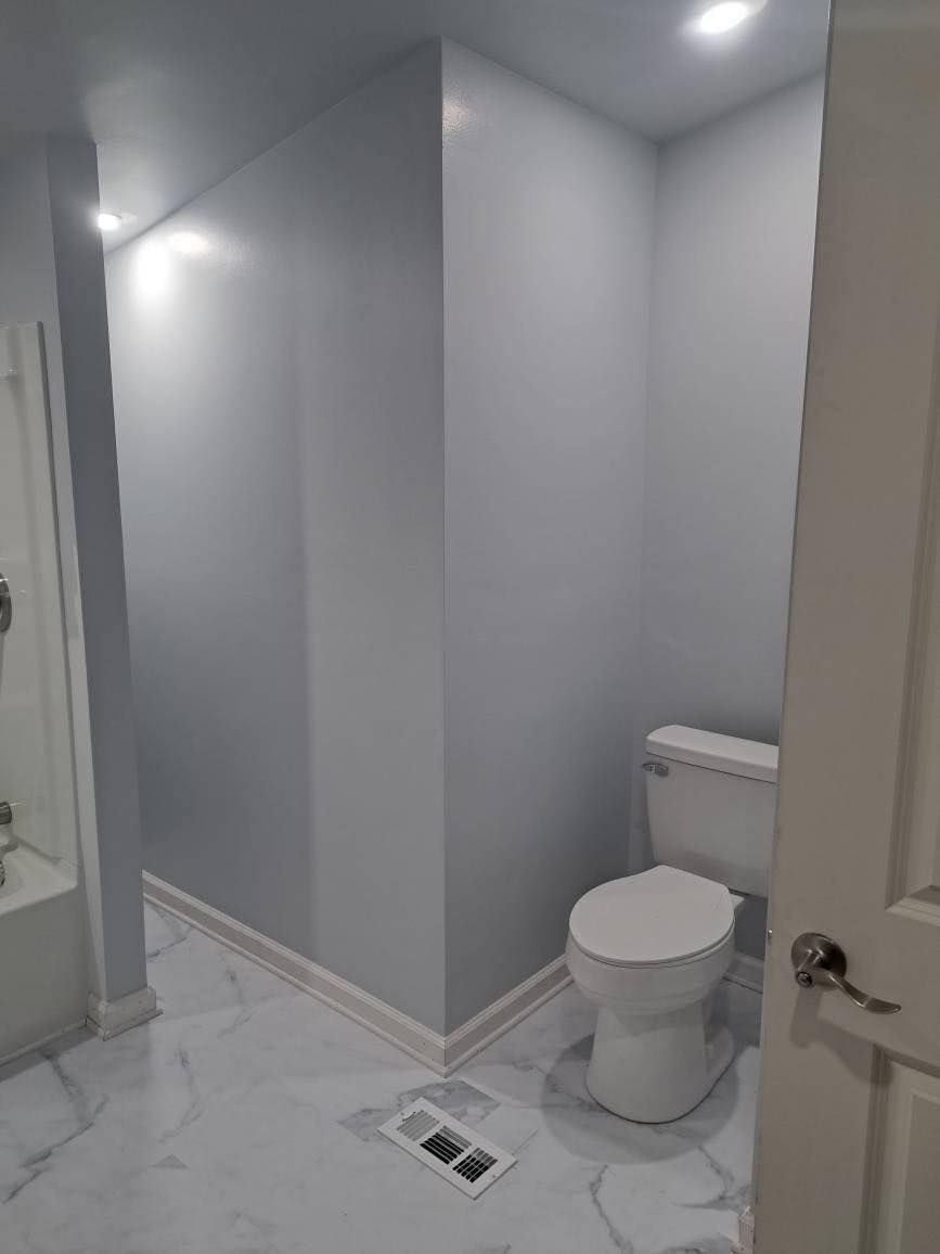 A light blue bathroom with white toilet, white door, and marble-look floor tiles. Recessed lighting.