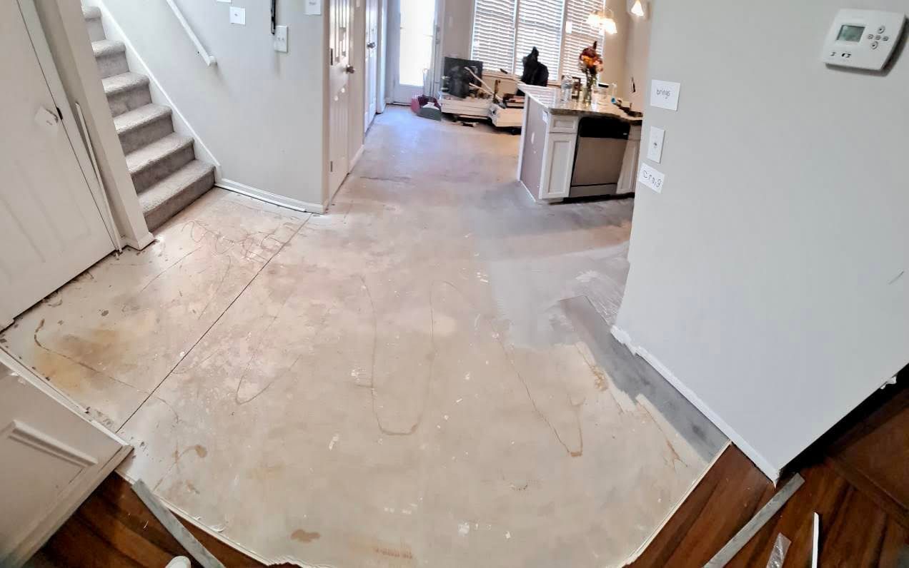 Interior view of a home with flooring removed, showing subfloor. Kitchen island, stairs, and doorway visible.