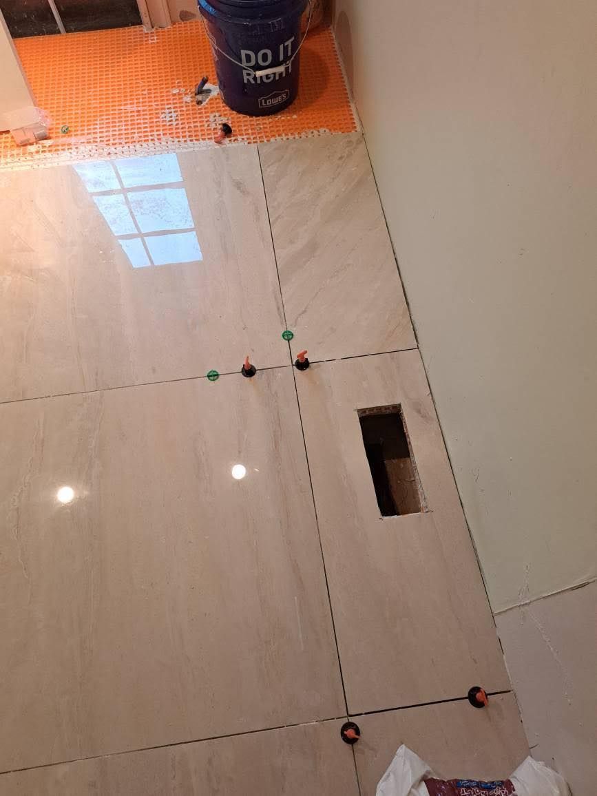 Bathroom floor under construction with large tiles, spacers, and a bucket. Beige tiles and an unfinished wall.