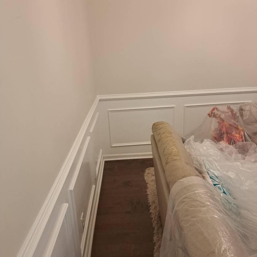 Corner of a room with white wainscoting and dark wood floor, a rolled-up rug is wrapped in plastic.