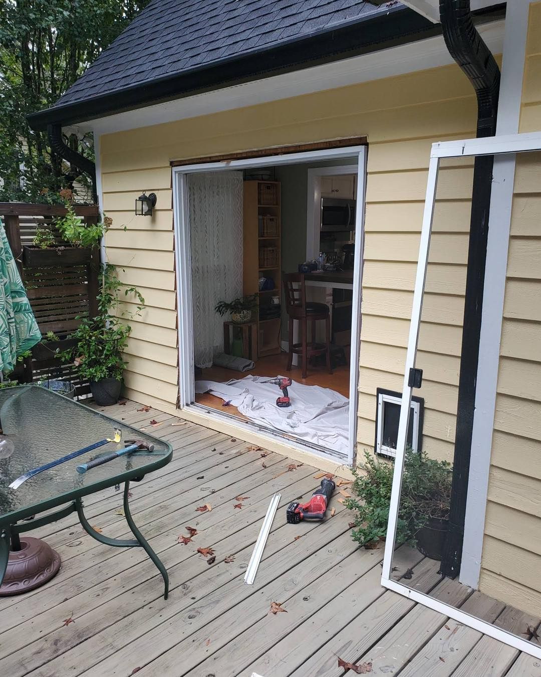 Sliding glass door repair on yellow house, with screen frame removed. Deck in foreground, plants visible.