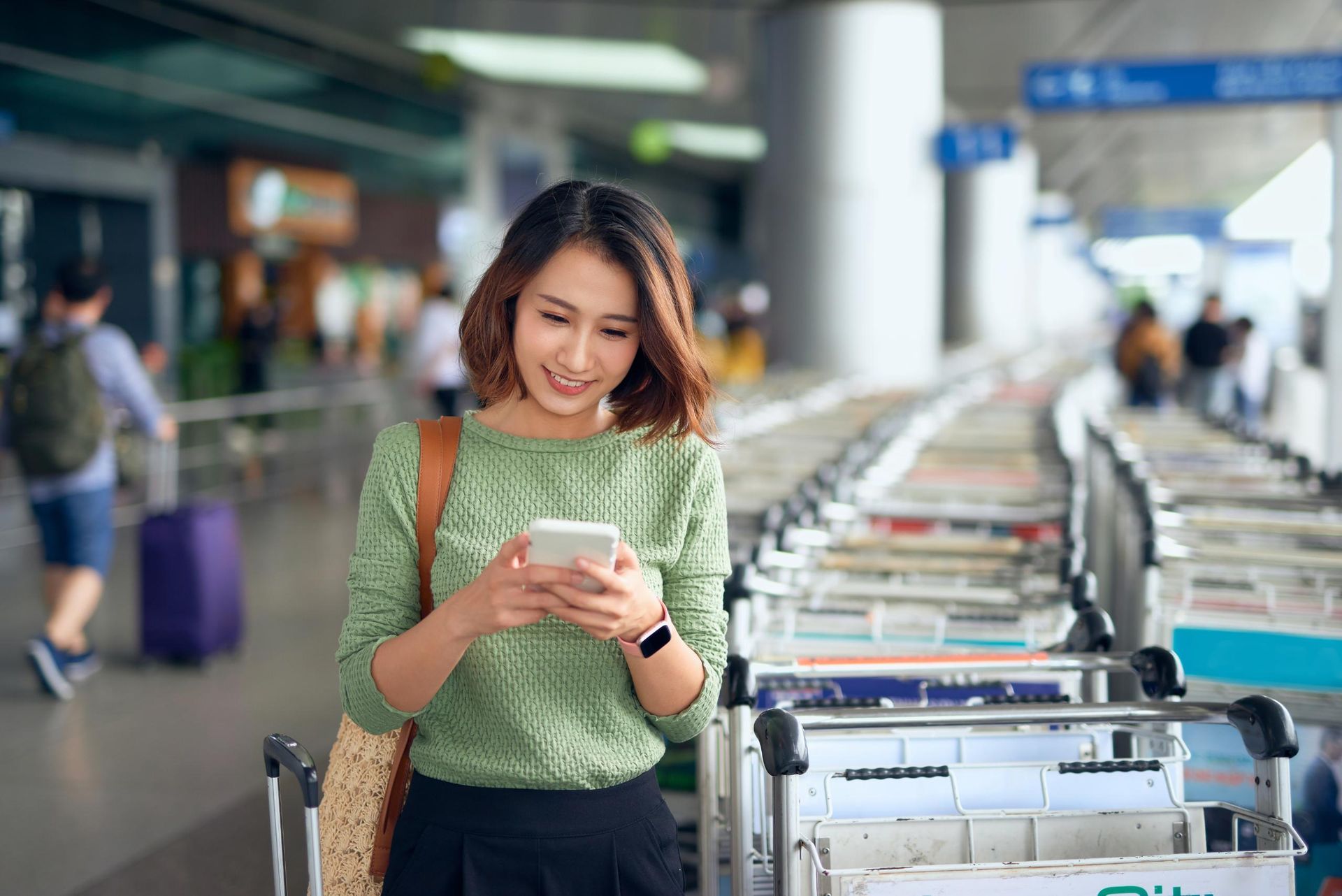 Woman in airport looking at phone, smiling, near luggage carts.