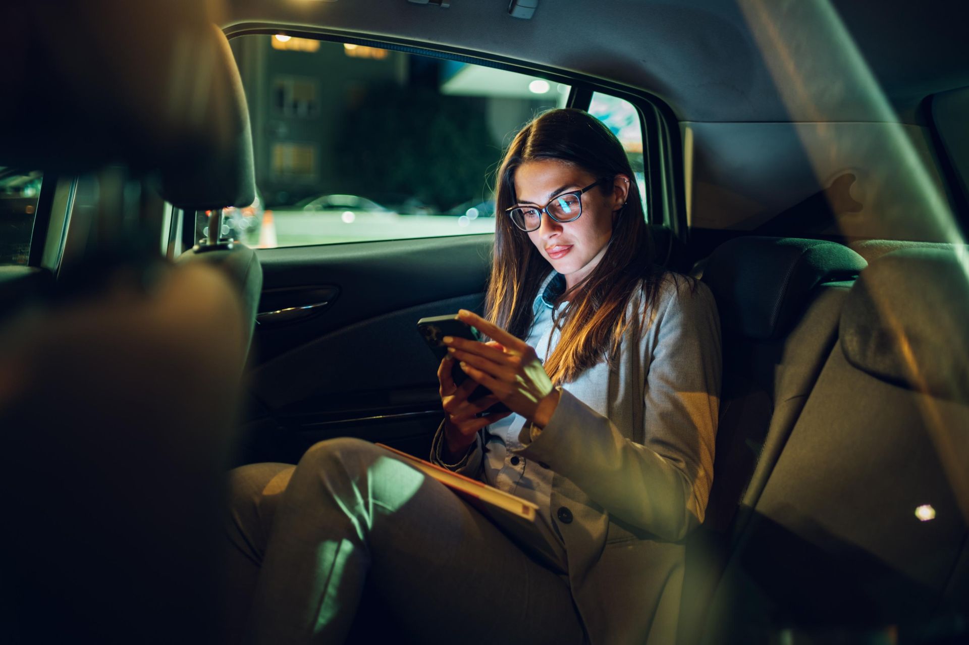 Woman in glasses, jacket, using phone in car at night.
