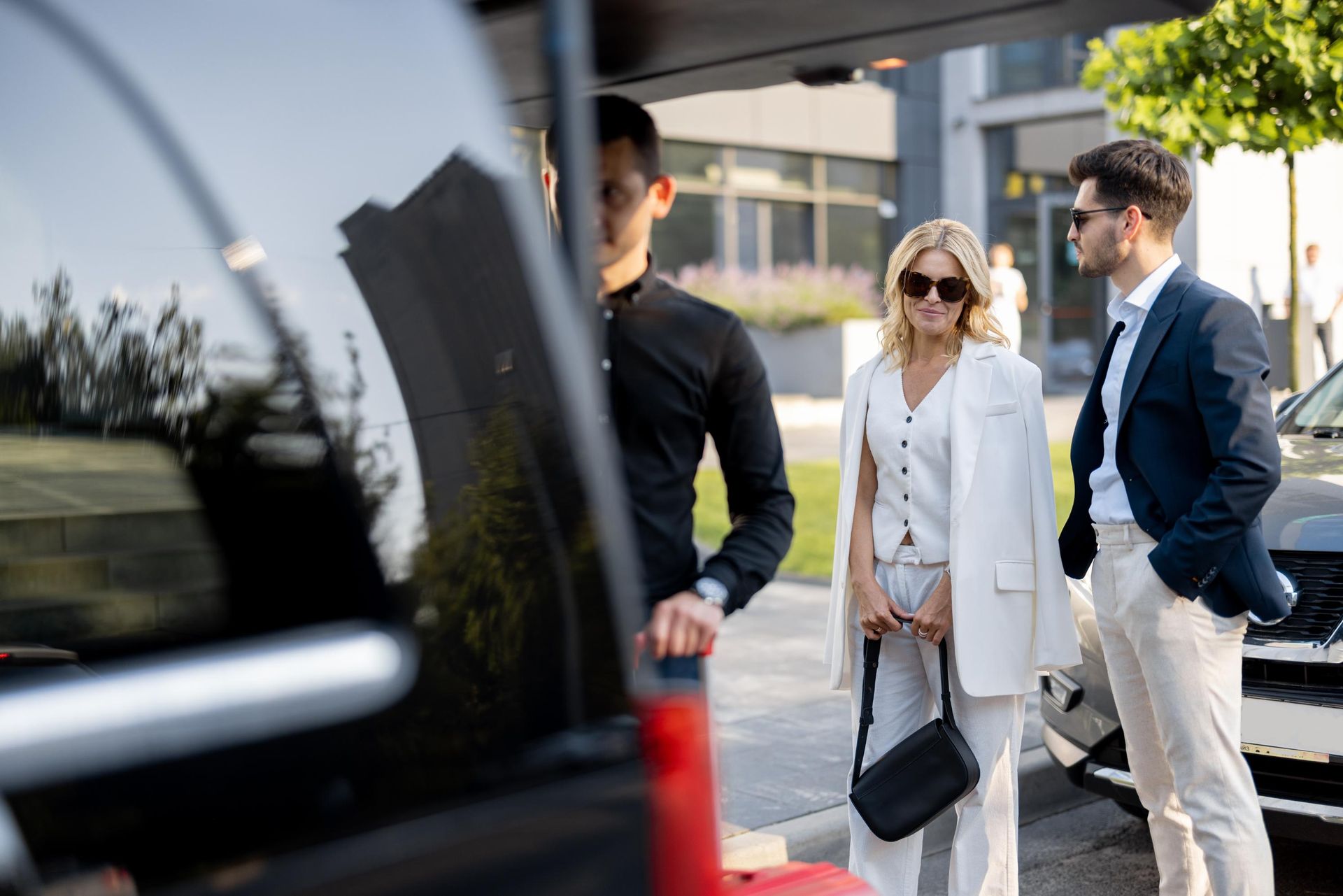 Man opening car trunk for well-dressed couple near modern building.