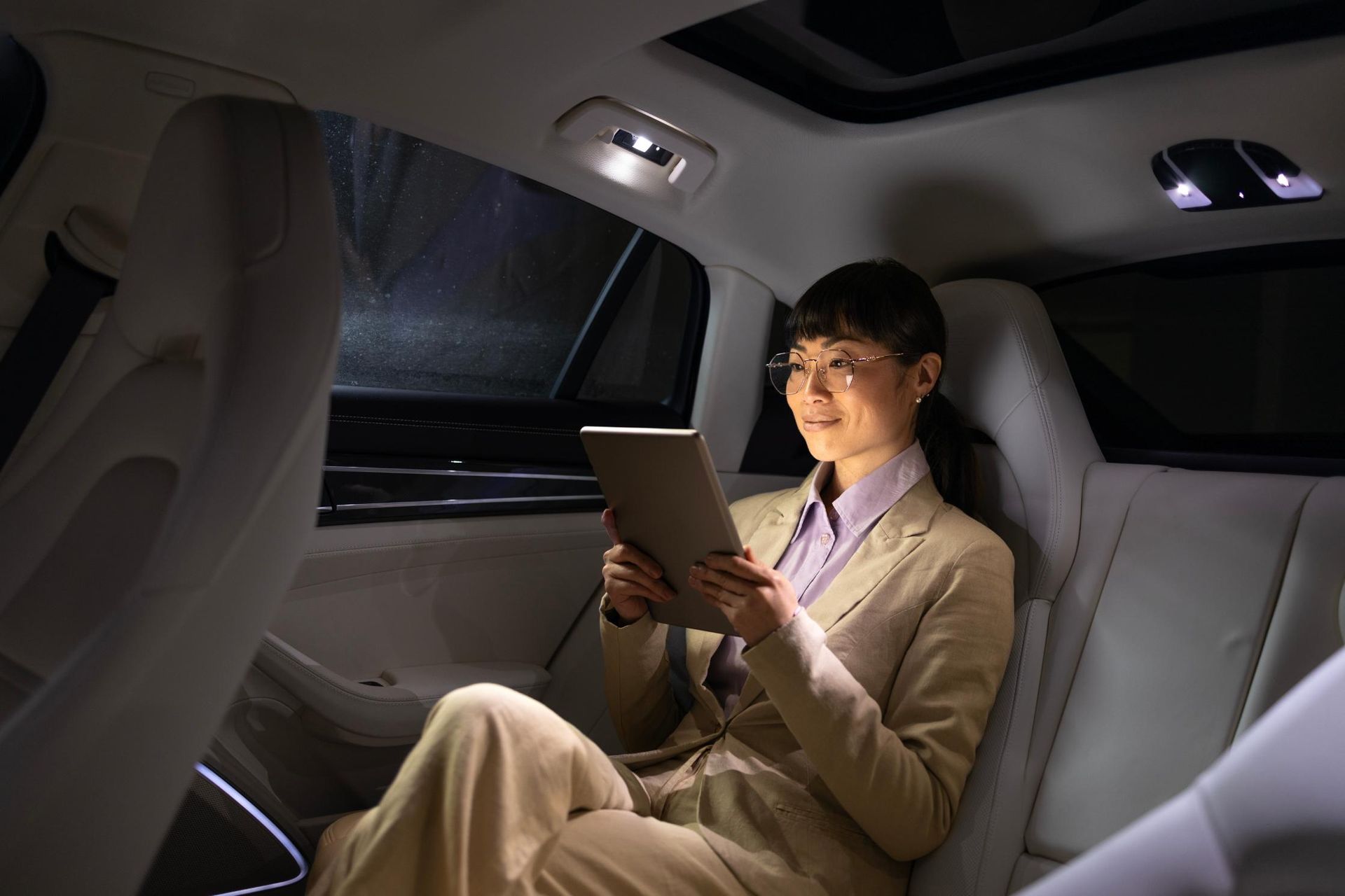 Woman in suit using a tablet in the backseat of a car. Lit interior, dark outside.
