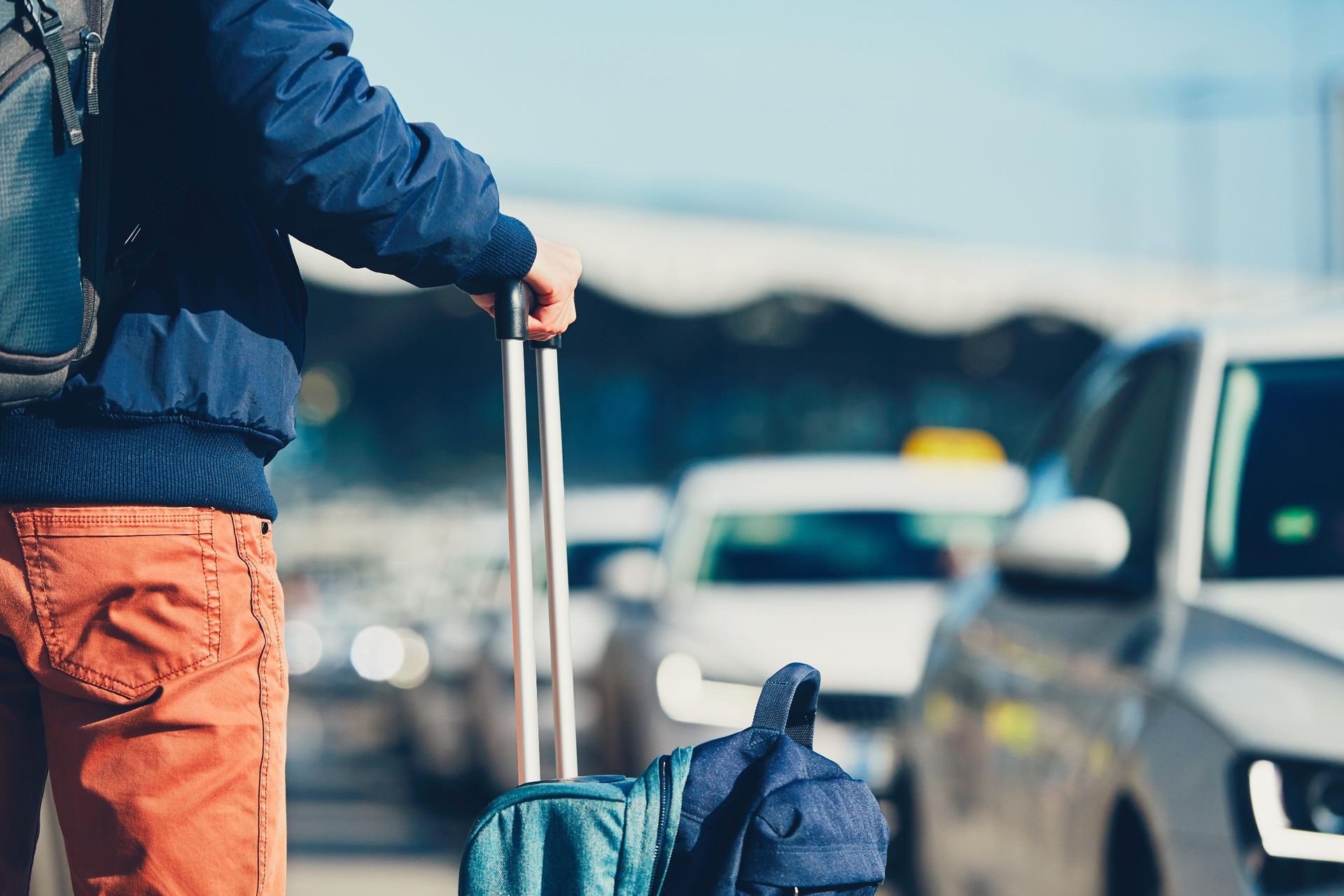 Person with backpack and rolling suitcase, standing near cars at an airport.