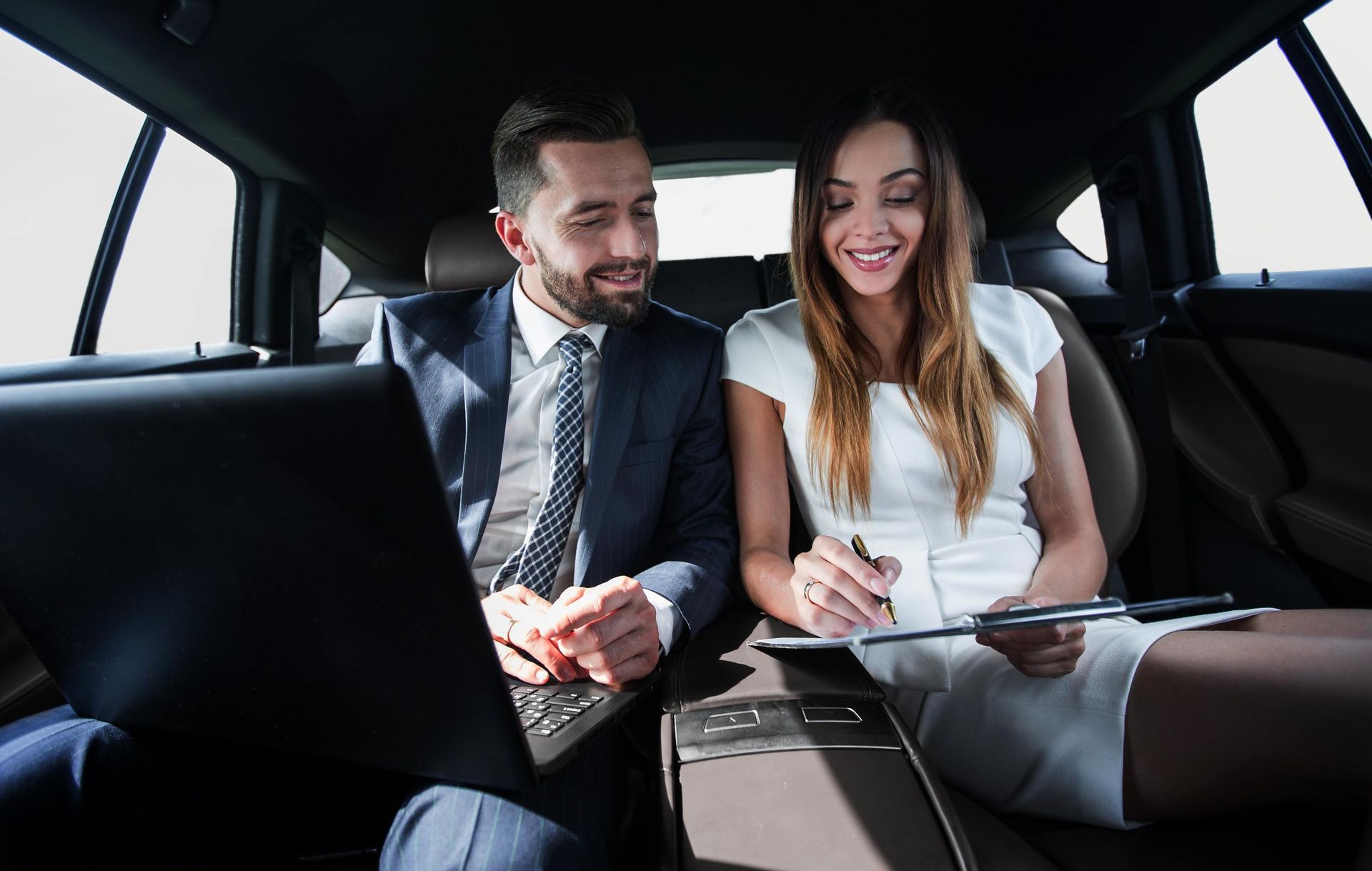 Man and woman in car, working. Man with laptop, woman with clipboard, both smiling.