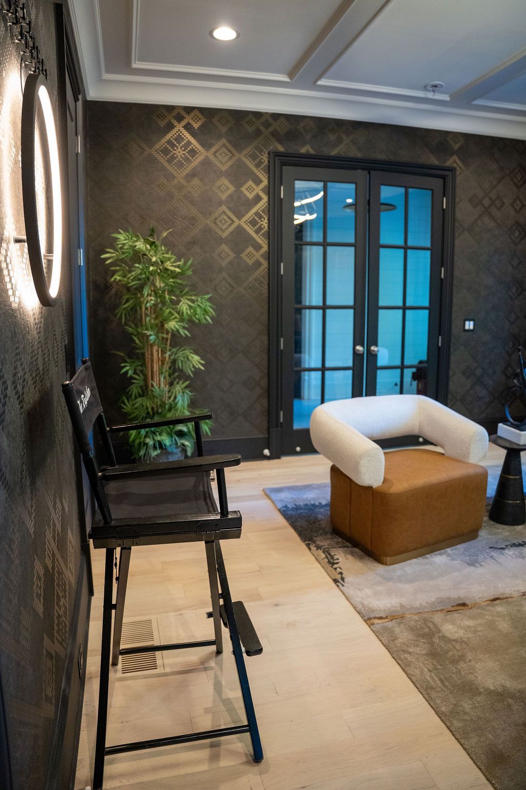 Black director's chair by a wall with gold geometric wallpaper, a white and brown armchair, and french doors.
