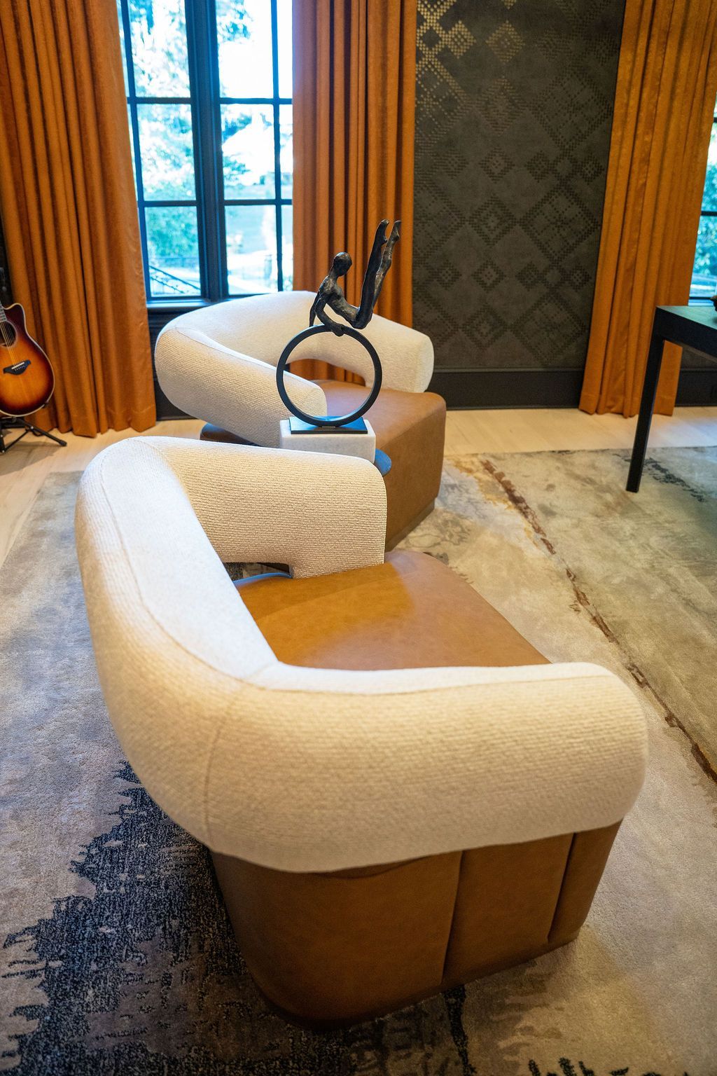 Two curved cream and tan chairs with a sculpture, set in a room with orange curtains and a patterned rug.