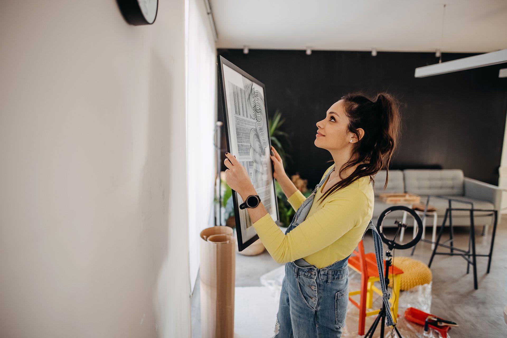Woman hangs a framed artwork on a white wall. She smiles, wearing a yellow sweater and denim overalls.