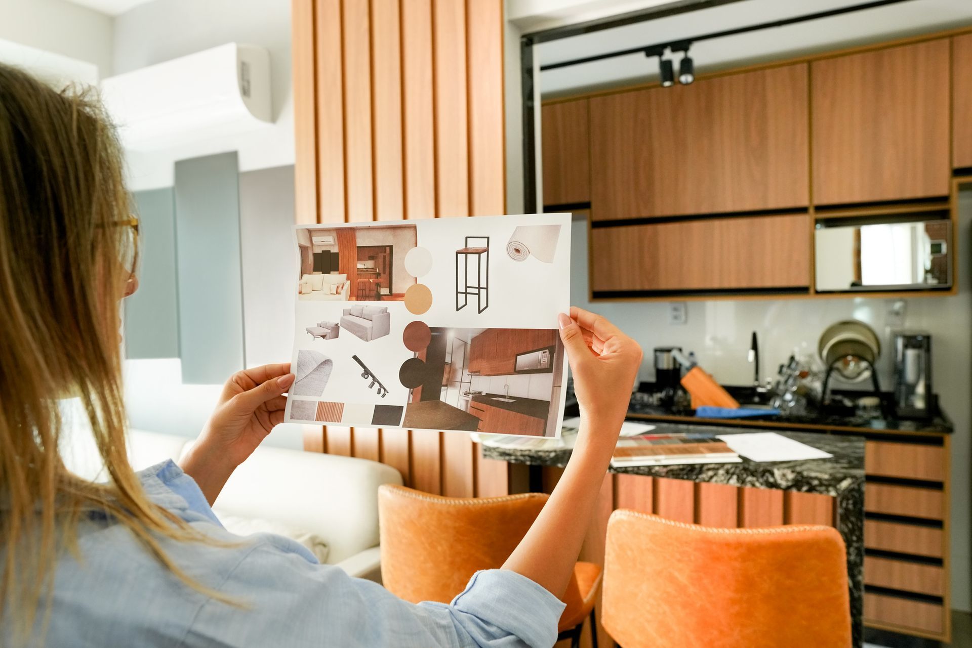 Woman holding design ideas, likely for kitchen renovation, with orange chairs and wooden accents visible.