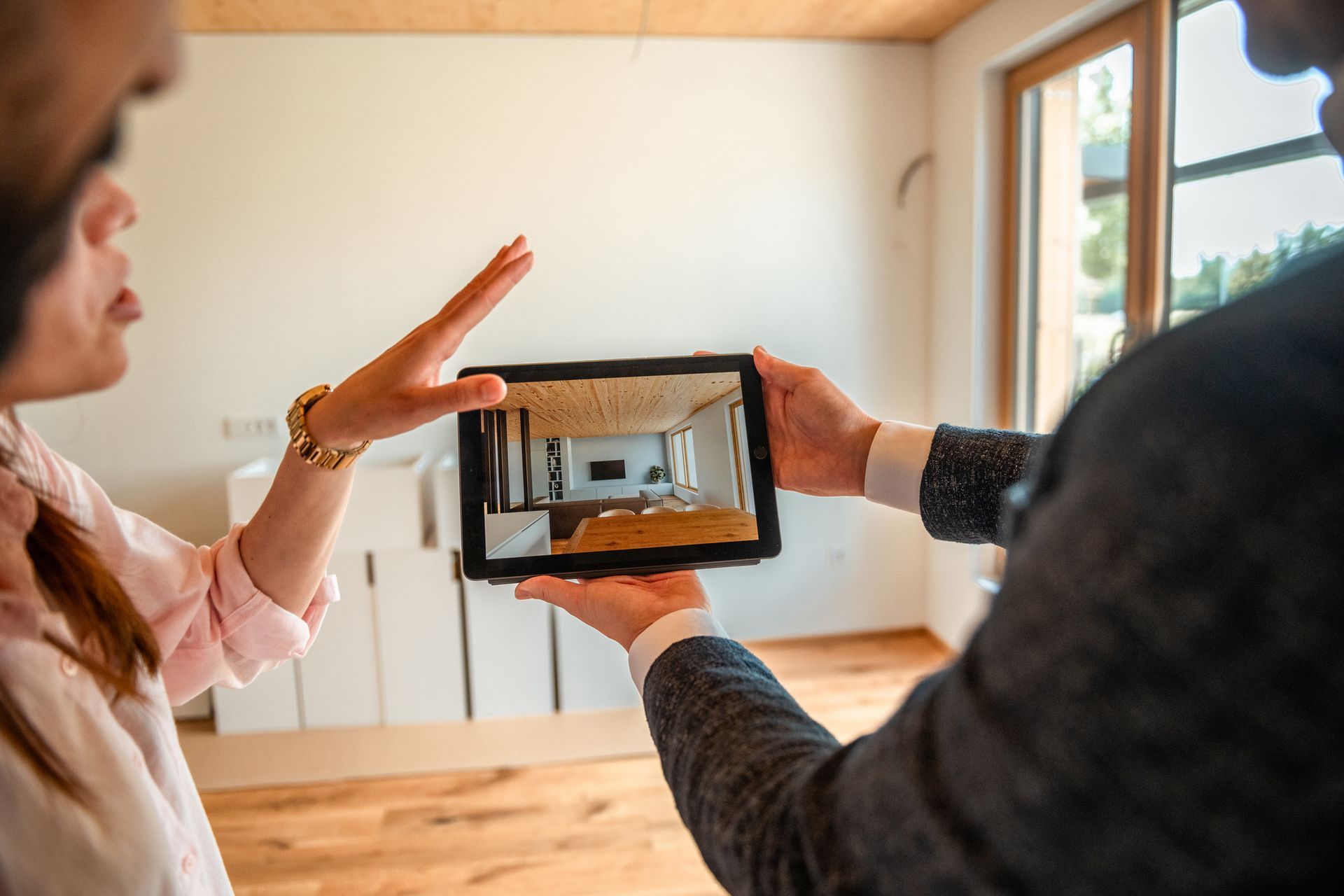 Two people looking at a tablet displaying a 3D rendering of a room. One gestures with a hand in front of the screen.