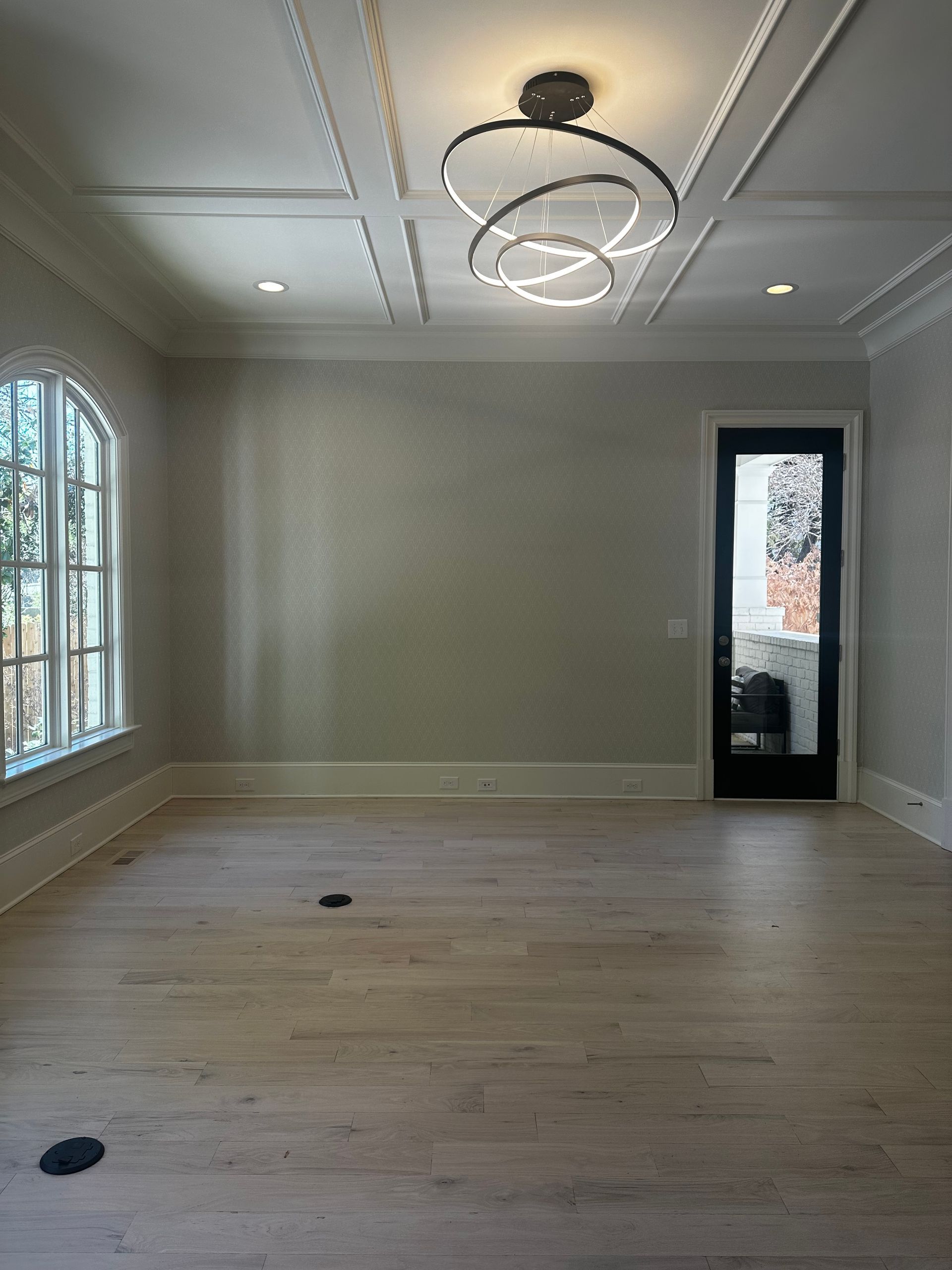 Empty room with wood floors, recessed lighting, and modern chandelier. Door and arched window visible.