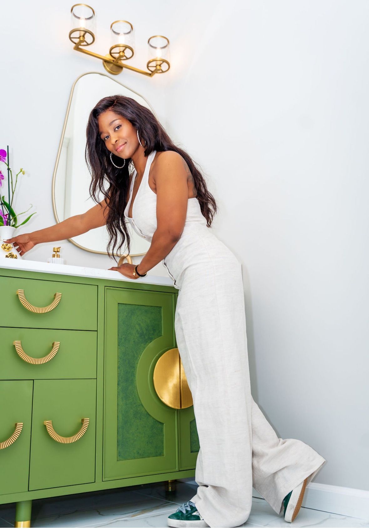Woman in white jumpsuit at a green vanity in bathroom, smiling.