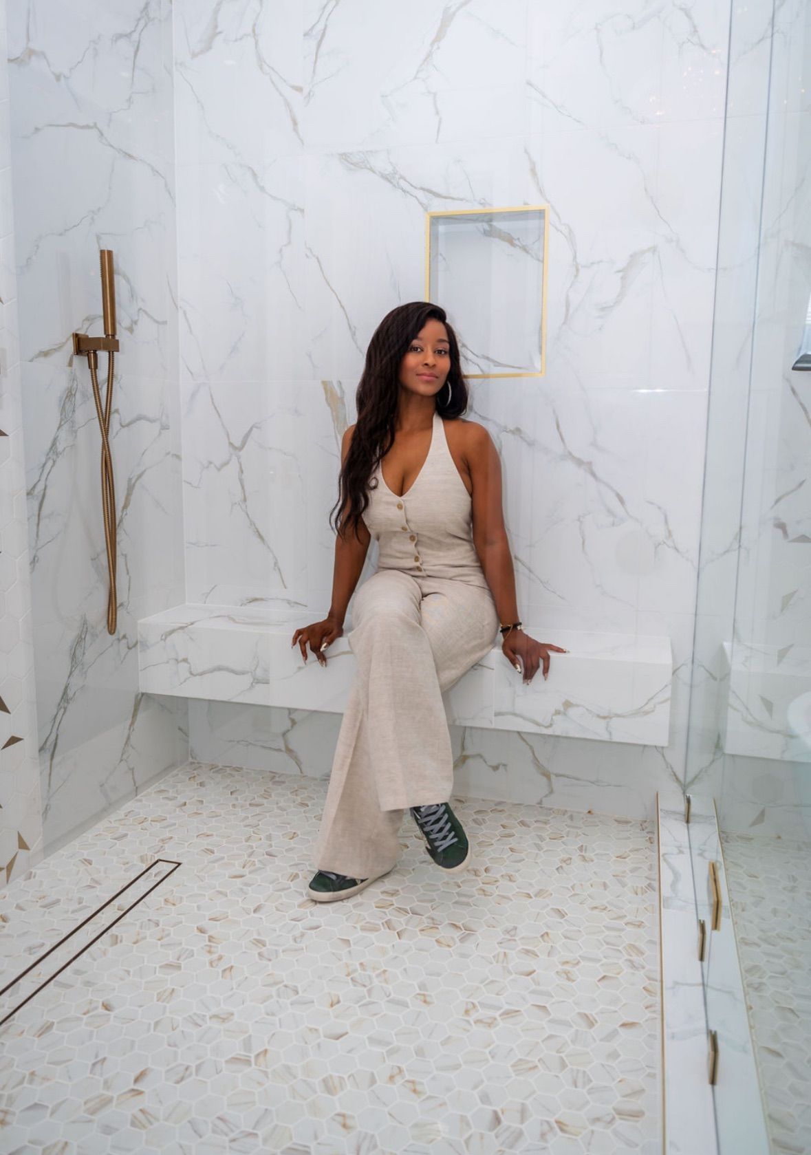 Woman in cream-colored outfit sits on a bench in a marble-tiled shower. She has dark hair and a slight smile.