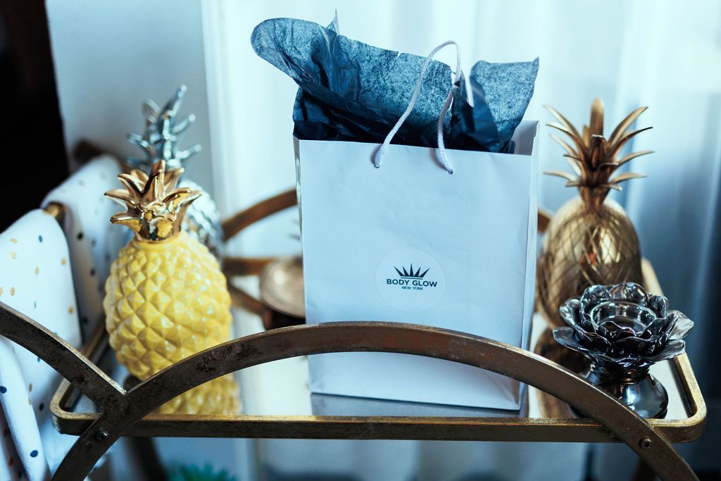 A white gift bag with blue tissue paper sits on a gold bar cart between two decorative gold pineapple figurines.