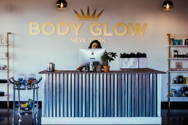 A woman is sitting at a desk in front of a body glow new york sign.