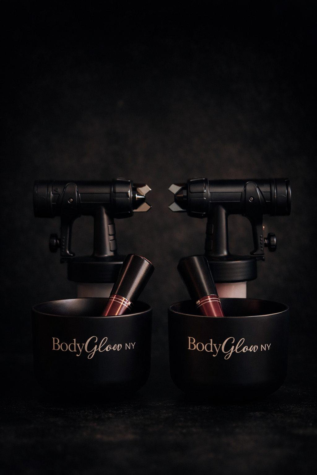 Two black spray tanning guns sit atop BodyGlow containers with lipstick inside. Dark background.