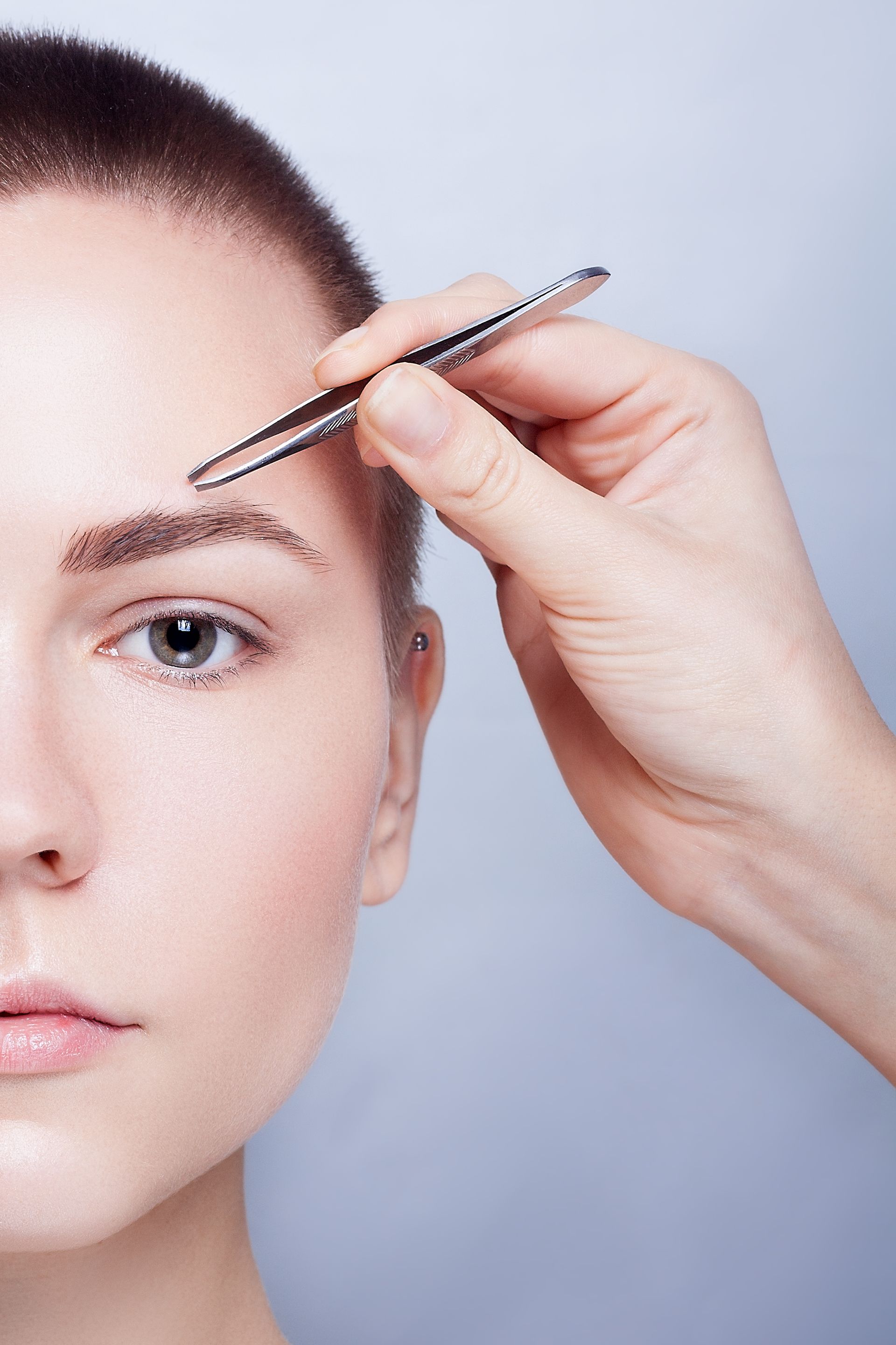 A woman with a shaved head is getting her eyebrows trimmed with tweezers.